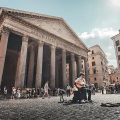 Un musicien joue de la guitare devant le Panthéon d'Agrippa, un monument historique de Rome, tandis que des touristes se promènent sur la place pavée.