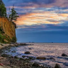 Zachód słońca nad morzem w Polsce. Klif porośnięty drzewami, kamienista plaża i statki na horyzoncie.