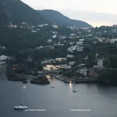 Vittoria Colonna a Ischia: case aggrappate alla collina, luci soffuse e barche a vela nella baia al crepuscolo.