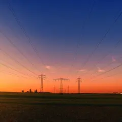 Linie energetyczne biegnące nad polem przy zachodzie słońca. W oddali widać przemysłowy krajobraz, przypominający o opłacie sieciowej stałej.