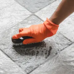 Mano con guanto arancione pulisce le fughe del pavimento con una spazzola, creando schiuma.