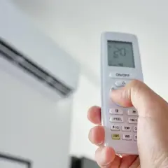 Mano sujetando un control remoto para encender aire acondicionado. La unidad interior está en el fondo.