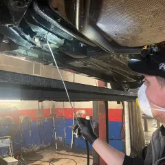Mechanik w masce i rękawiczkach aplikuje środek konserwujący na podwozie samochodu. Długi aplikator pozwala dotrzeć do trudno dostępnych miejsc, co jest kluczowe, by wiedzieć, ile trwa konserwacja podwozia.