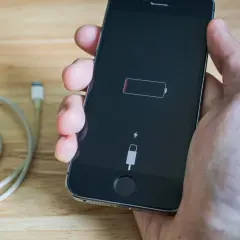 Dłoń trzyma iPhone'a z ikoną pustej baterii i symbolem błyskawicy, co oznacza, że iphone nie ładuje się. Obok leży kabel.