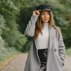 Elegancka kobieta w szarym płaszczu męskim, białym golfie i czarnej spódnicy, poprawia czapkę. Stylowa stylizacja na jesienny spacer.