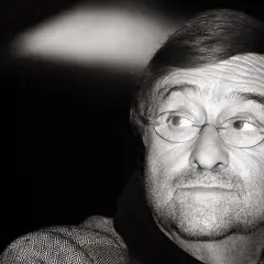 Ritratto in bianco e nero di un uomo con occhiali rotondi, barba e un'espressione pensierosa. Il suo sguardo è rivolto di lato, come se stesse ascoltando attentamente il **caruso testo**.