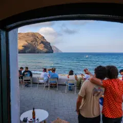 Dos amigos disfrutan de la vista al mar, con gente sentada en mesas. El manteca las negras, un día perfecto.