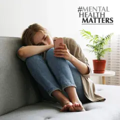 Junge Frau sitzt allein auf Sofa, depressiv weil single. Sie hält ihr Handy, #MENTAL HEALTH MATTERS.