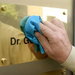 Hand reinigt Messing-Namensschild mit Tuch. Der Schriftzug "Dr. G." wird sichtbar.