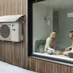 Unidad exterior de aire acondicionado Daikin instalada con la distancia adecuada a la pared de madera. Dentro, una pareja juega.
