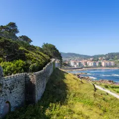 A historic stone wall winds along the coast of Baiona, Portugal, overlooking the blue sea and a town nestled on the hills.