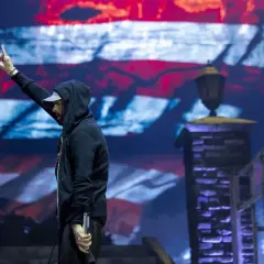 Eminem na koncercie w Polsce, z ręką uniesioną w geście pozdrowienia, na tle ekranu z amerykańską flagą.