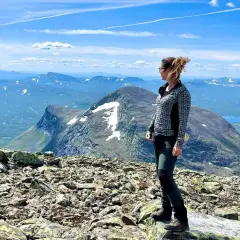 En kvinna står på en stenig topp med utsikt över ett vidsträckt fjällandskap. En vy som liknar den från en **webbkamera Helags**.