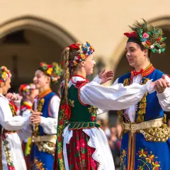 Młoda para w barwnych strojach ludowych tańczy krakowiaka. Dziewczyna w wianku z kwiatów i chłopak w ozdobnej czapce z piórami.