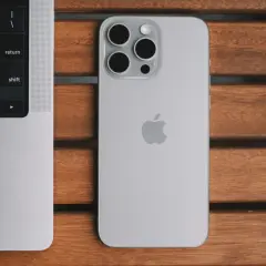 Srebrny iPhone 13 Pro z systemem iOS 26.4 obok klawiatury MacBook'a na drewnianym stole.