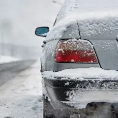 Samochód pokryty śniegiem na poboczu drogi. Widać dym z rury wydechowej, co sugeruje zdalne uruchamianie silnika.