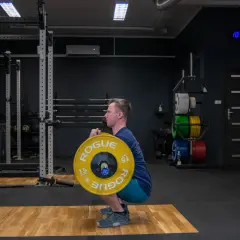Mężczyzna wykonuje zercher squat z ciężarem w zgięciu łokci. Siłownia z wolnymi ciężarami i sprzętem do treningu siłowego.