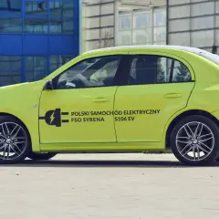 Jasnozielony FSO Syrena S106 EV, jeden z polskich samochodów elektrycznych, parkuje przed nowoczesnym budynkiem.