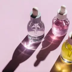 Trzy buteleczki z pipetką: przezroczysta, różowa i żółta. Cienie i refleksy światła na różowym tle. Czy zawierają parabeny w kosmetykach?
