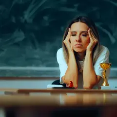 Lehrerin mit Händen an den Schläfen, Augen geschlossen, wirkt erschöpft. Ein Pokal und Bücher auf dem Tisch deuten auf den Schulalltag hin, der zu Burnout bei Lehrern führen kann.