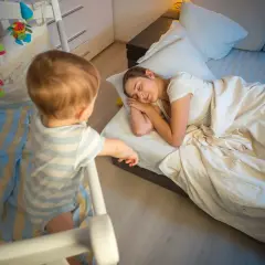 Un neonato di 7 mesi, in piedi nella sua culla, osserva la mamma addormentata nel letto.