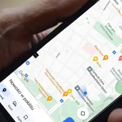 Dłoń trzyma telefon z mapą Google. Widoczna jest opcja udostępnianie lokalizacji, a także pobliskie restauracje i sklepy.