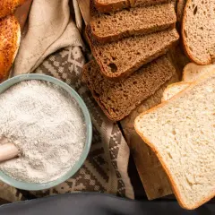 Mąka w misce, kromki chleba i obwarzanek. W tych produktach jest gluten, który jest podstawą wypieków.