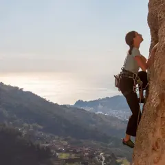 Eine Frau klettert an einer Felswand in Finale Ligure. Im Hintergrund glitzert das Meer.