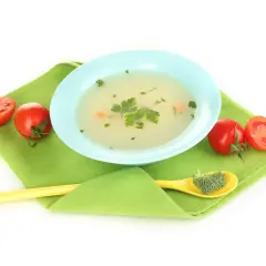 Brodo vegetale neonati in ciotola azzurra, con pomodorini e un cucchiaino giallo con broccoli.