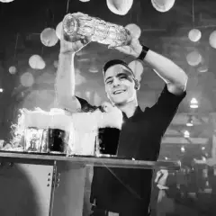 Uśmiechnięty barman z shakerem nad głową, przygotowuje drinki z dymem.