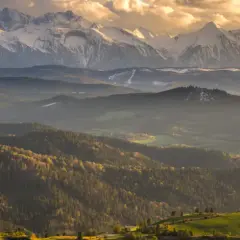 Majestatyczne szczyty górskie w słońcu, otoczone łagodnymi wzgórzami i lasami.