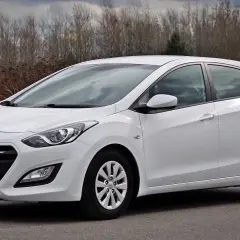 Biały Hyundai hatchback zaparkowany na drodze, z zaroślami w tle.