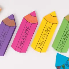 Bunte Stift-Bastelvorlagen mit "Einladung" beschriftet. Eine kleine Tafel mit ABC und eine blaue Wimpelkette sind ebenfalls zu sehen.