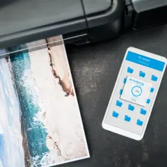Jak wydrukowac cos z iPhone? Widok telefonu z ekranem "Printing 38%" obok drukarki drukującej zdjęcie plaży.
