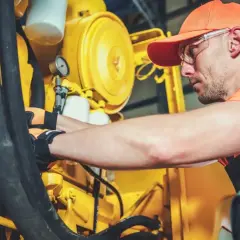 Técnico con guantes y gafas realizando la limpieza de maquinaria industrial amarilla, revisando mangueras y componentes.
