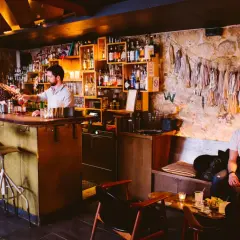 Ambiance feutrée dans ce bar caché. Un barman sert un cocktail, des clients discutent au comptoir et sur une banquette.