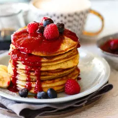 Ein Stapel goldbrauner Dinkel Pancakes, übergossen mit roter Beerensauce und garniert mit frischen Beeren und Bananenscheiben.