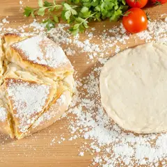 Une recette de pain maison : une miche dorée saupoudrée de farine à côté d'une pâte à pain crue, avec des tomates et du persil en arrière-plan.