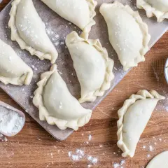 Surowe pierogi ruskie ułożone na desce, gotowe do zamrożenia. Obok miseczka z farszem i drewniana łopatka z mąką.