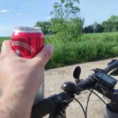 Rowerzysta trzyma puszkę napoju, planując jazdę rowerem po alkoholu. W tle zielone pola i błękitne niebo.