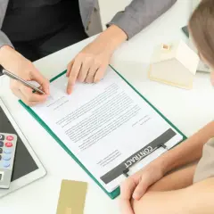Kobieta podpisuje umowę kredytową. W tle model domu, kalkulator i karta. Wspólny kredyt bez ślubu a rozstanie – ważne są warunki umowy.