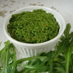 Pesto z rukoli w białej miseczce, otoczone świeżymi liśćmi rukoli. Idealne do makaronu lub jako dodatek do kanapek.