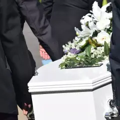 Pogrzeb członka rodziny, który nie był ubezpieczony. Biała trumna niesiona przez żałobników, ozdobiona bukietem białych kwiatów. Zasiłek pogrzebowy na nieubezpieczonego członka rodziny.