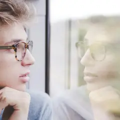 Młody mężczyzna w okularach patrzy w okno, jego odbicie sugeruje głęboką refleksję i inteligencję intrapersonalną.