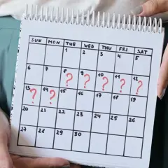 Kalender mit roten Fragezeichen, die auf unregelmäßige Zyklen und den Zusammenhang zwischen **Stress und Periode** hinweisen.