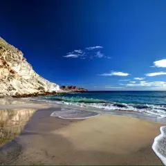 Playa de arena dorada y acantilados rocosos en la cala del plomo, con el mar azul intenso y olas espumosas.