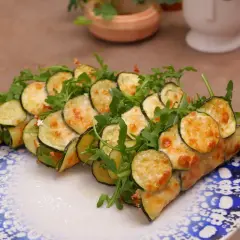 Rotolini di zucchine e rucola, cotti in padella e gratinati con formaggio fuso, serviti su un piatto decorato.