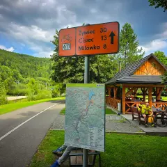 Znak szlaku rowerowego Velo Sola z odległościami do Żywca, Ciśca i Milówki. Obok mapa i wiata.