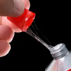Main d'une personne tenant le bouchon rouge d'un tube de colle super glue, avec des fils de colle s'étirant. Attention, colle super glue sur les doigts dangereux.
