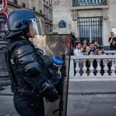 Policjant w pełnym rynsztunku ochronnym z tarczą, obserwuje tłum za barierką.